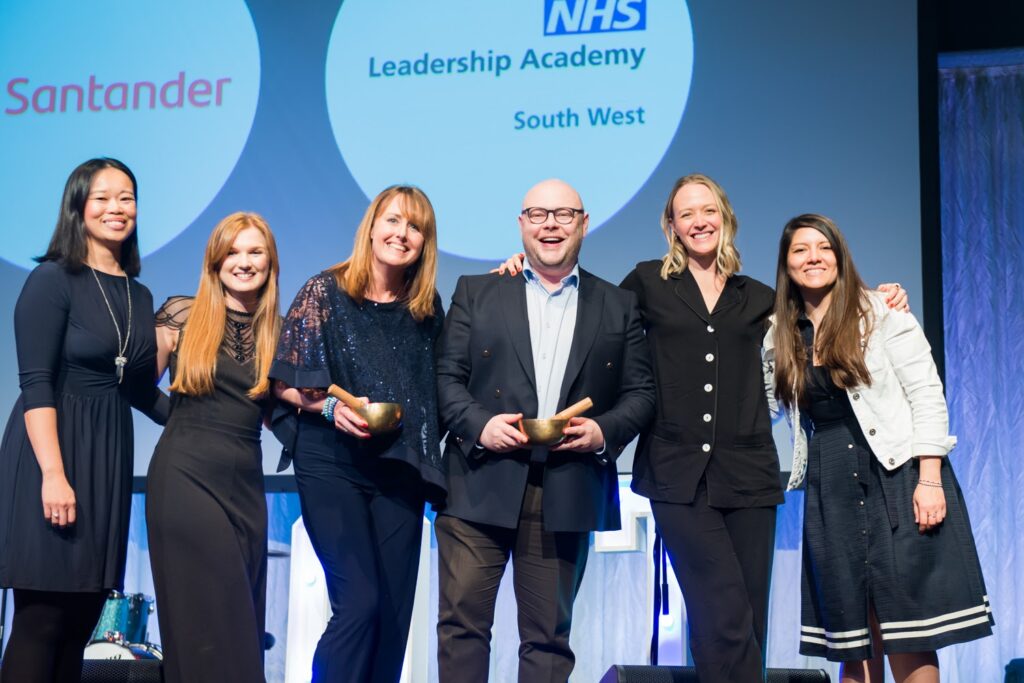 Receipt of award pictured: From left , On a stage in front of a screen that reads NHS Leadership Academy South West. With arms around each other in front of the screen, Leadership Development Coordinators Connie Cheung with shoulder length black hair, smiling and wearing a navy blue dress, Ellie Jullens, with waist-length red hair, smiling and wearing a black dress, Leadership Development  Senior Manager Zoe Spittle with shoulder length strawberry blonde hair, smiling and wearing a sparkly navy jumpsuit and Inclusion Coordinator Nora Latapi-Dean with waist-length brown hair smiling and wearing a white jacket and blue navy dress pictured.