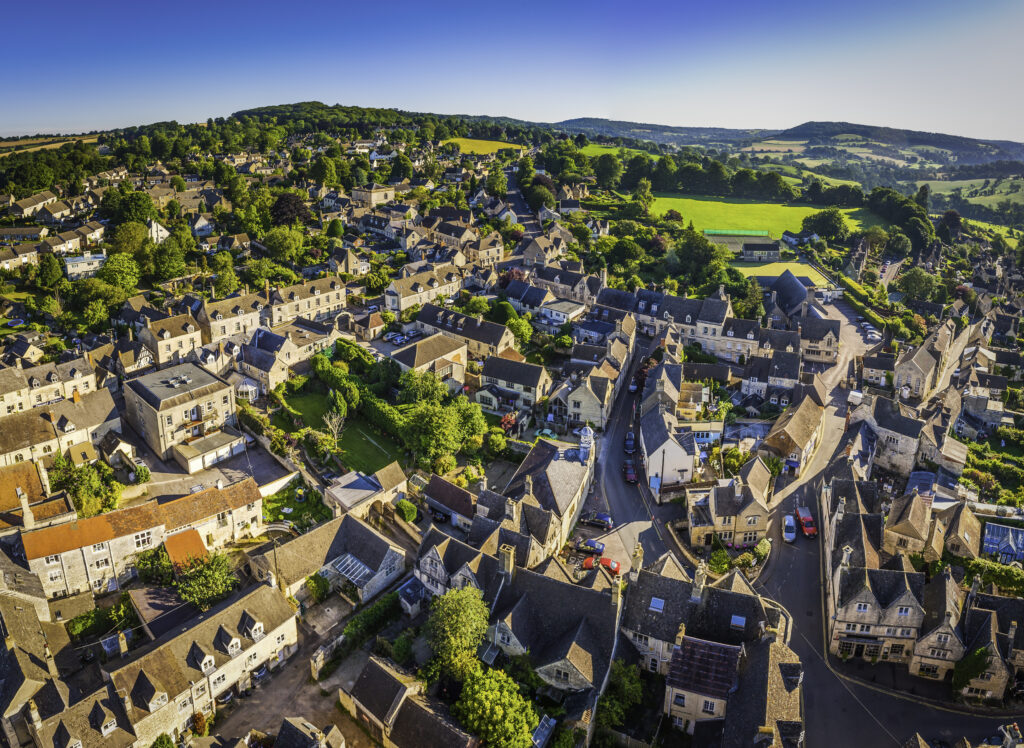 Aerial view over the iconic Cotswold village of Painswick, with its honey coloured limestone cottages deep in the bucolic countryside of Gloucestershire, UK, framed by vibrant green patchwork fields and clear blue summer skies. ProPhoto RGB profile for maximum color fidelity and gamut.